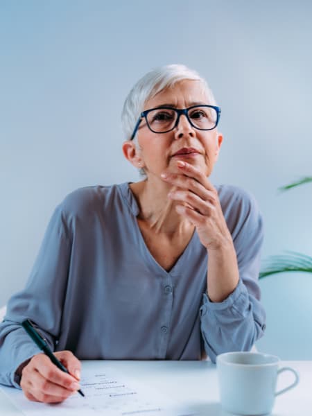 A senior woman thoughtfully gazing into the distance while filling out a paper | Think Clearly, Live Well. Perfect Synergy®.